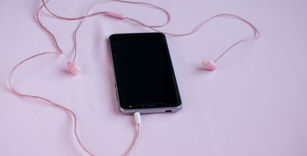An older smartphone sitting on a pale pink surface with pink wired headphones plugged in.