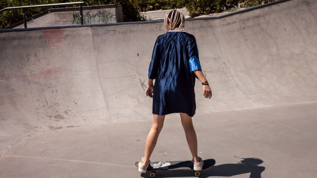 a young woman in an oversized shirt with bare legs and trainers riding a skateboard in a skate park.  Girl is facing away from the camera and has dyed blonde little braids in her hair.