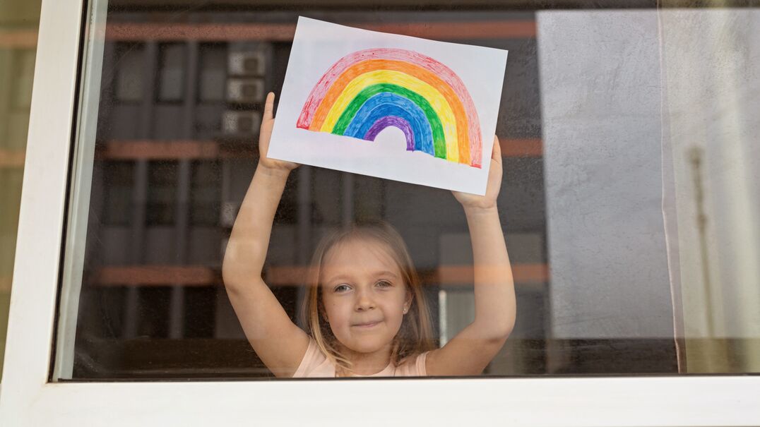 Young girl standing at a window holding up a rainbow drawing